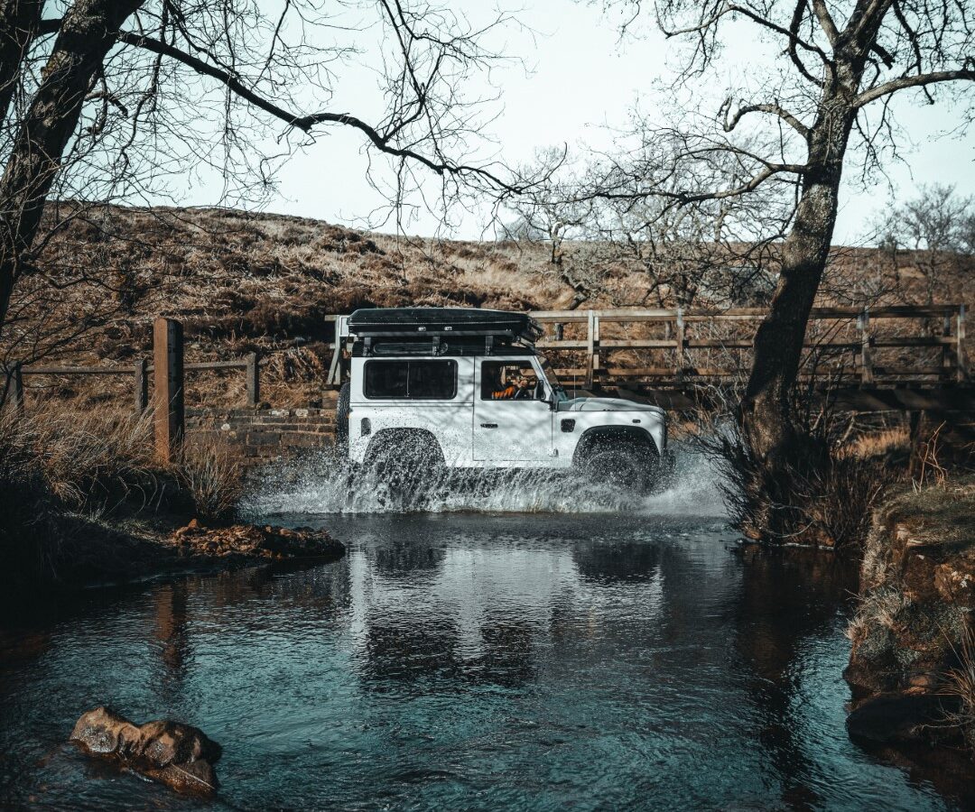 A white Defender off-road vehicle skillfully crosses a shallow stream, surrounded by leafless trees and a wooden fence—a perfect scene for an adventurous camping expedition.