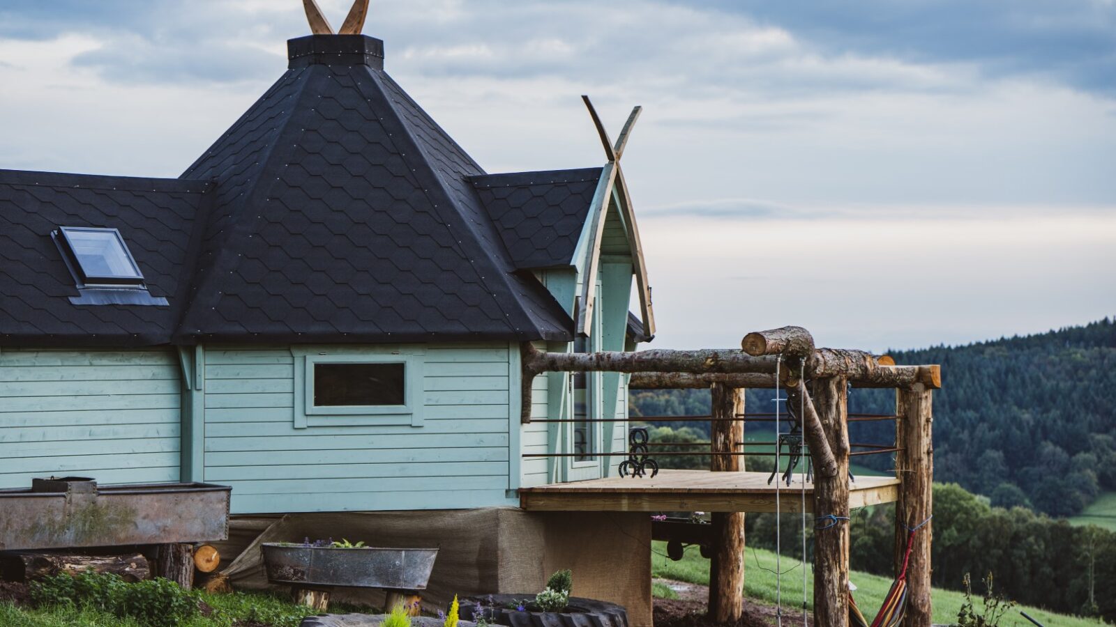 A wooden cabin with a unique roof design and a small deck, nestled against a green landscape under a cloudy sky, stands just beyond the mystical Dragons Gate.