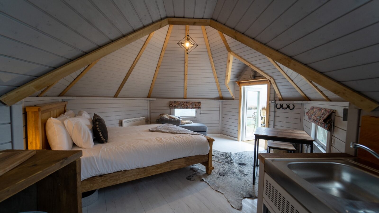 A cozy loft near Dragons Gate, featuring a wooden bed, white bedding, a small kitchenette, and natural light streaming through open doors and windows.