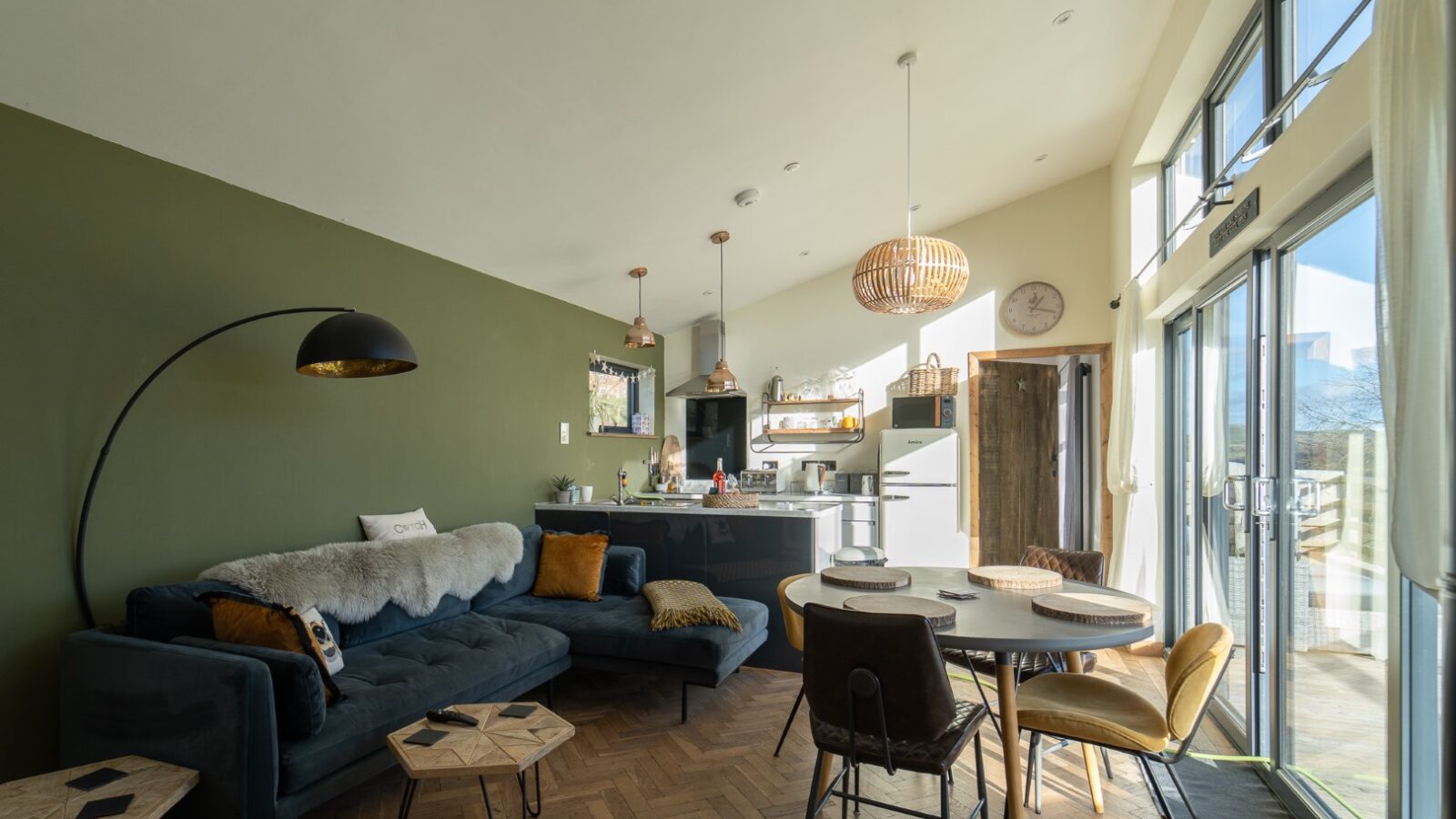 The Penlan Lodges offer a modern living room featuring a green accent wall, blue sofa, and round dining table seamlessly integrated with an open kitchen in a bright, spacious setting.