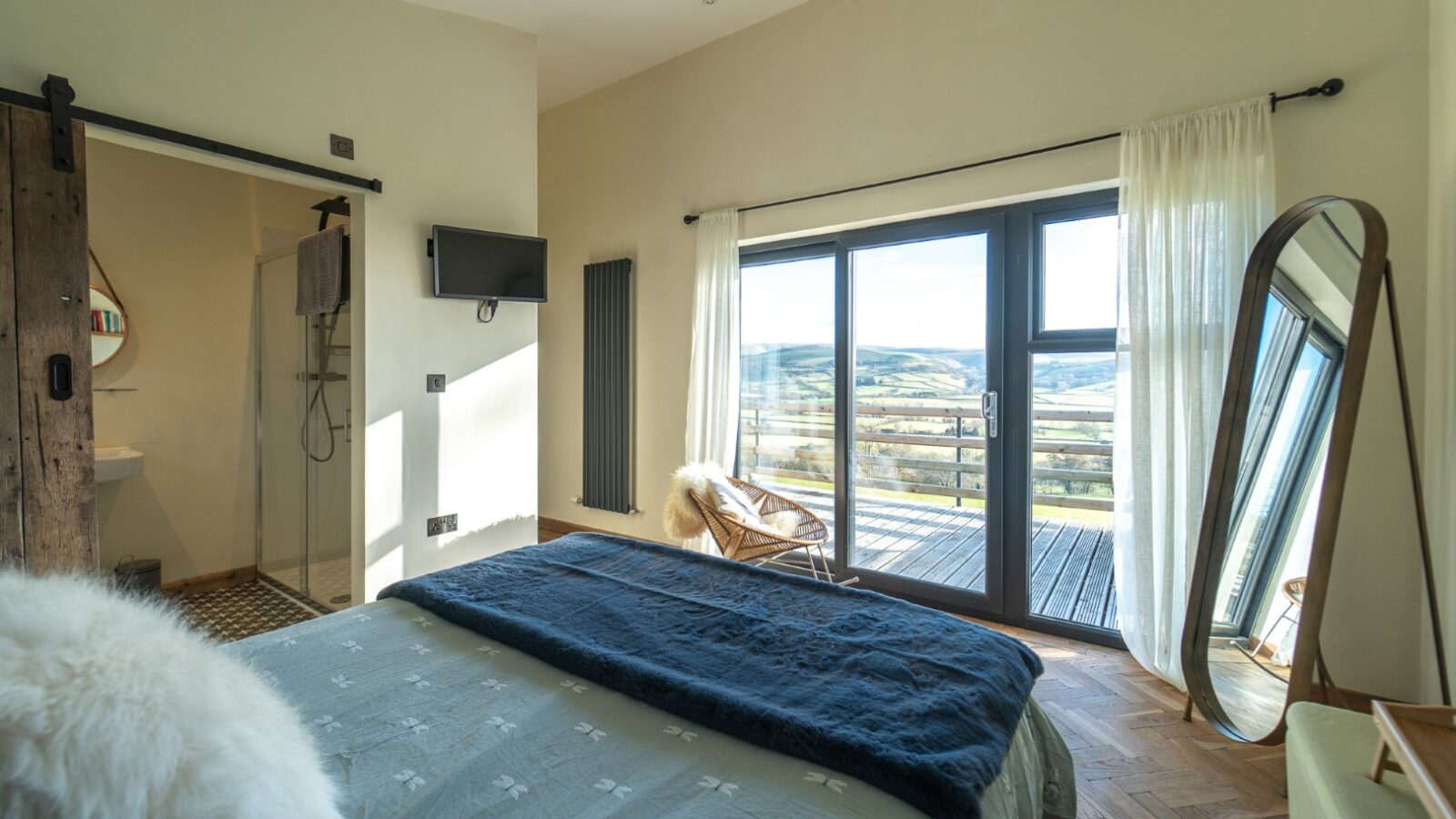 The Penlan bedroom features a cozy double bed, large mirror, wall-mounted TV, and a sliding door leading to a luxurious bathroom. Floor-to-ceiling windows in this stunning lodge offer breathtaking views of the scenic landscape beyond.