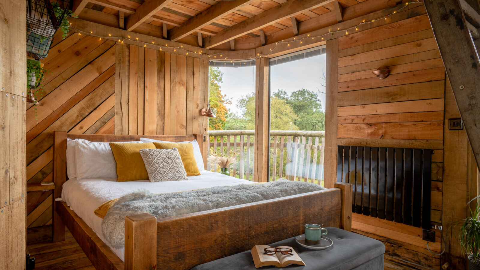 The cozy wooden bedroom, tucked within the Trawscwm Treehouse, features a large bed adorned with fairy lights and a window view of lush greenery. A book and mugs rest invitingly on a bench at the foot of the bed, creating the perfect spot for relaxation.