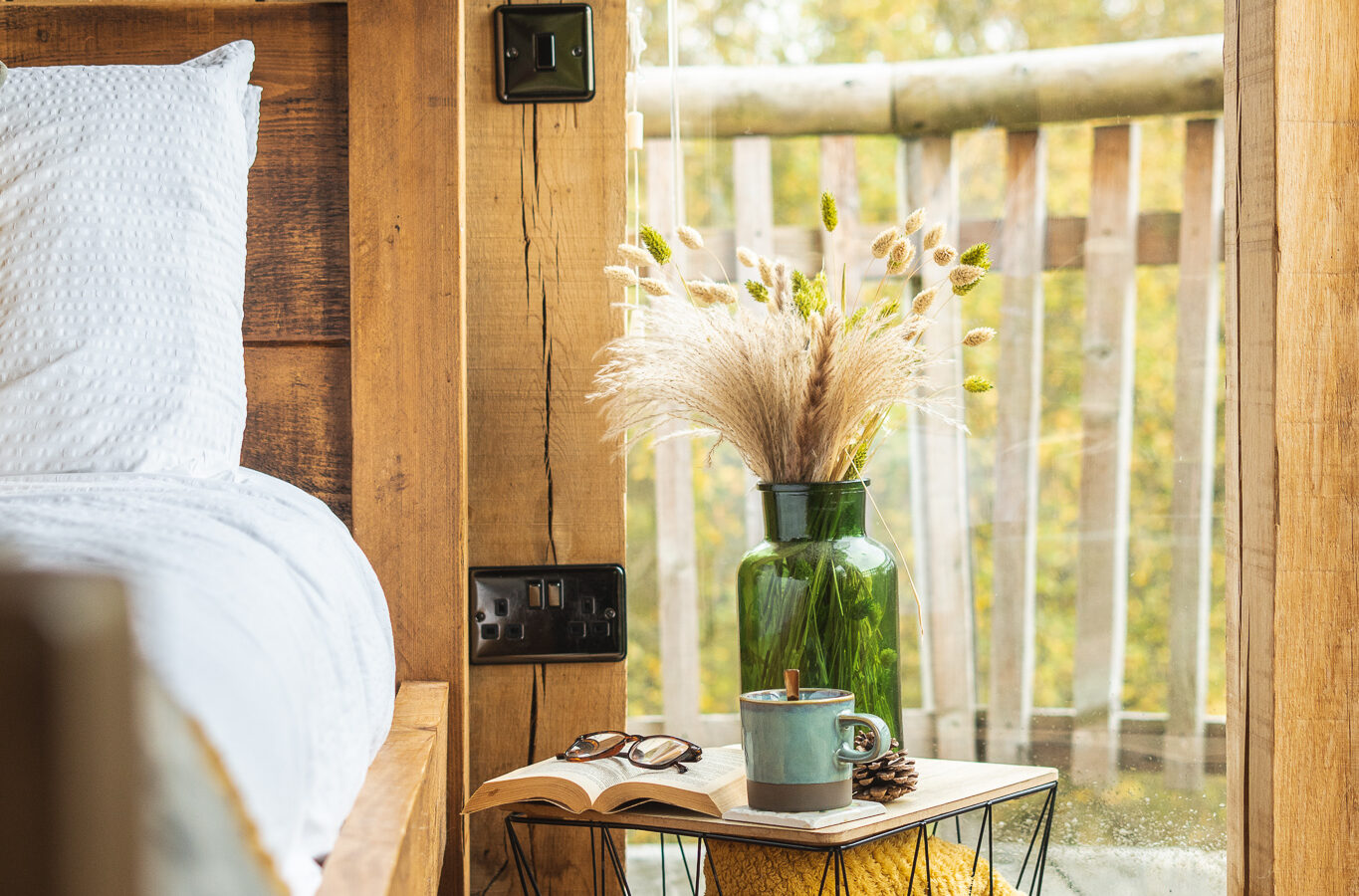 The cozy wooden bedroom in the Trawscwm Treehouse features a bed, a bedside table with a vase, a mug, and a book by the window overlooking nature's serene beauty.