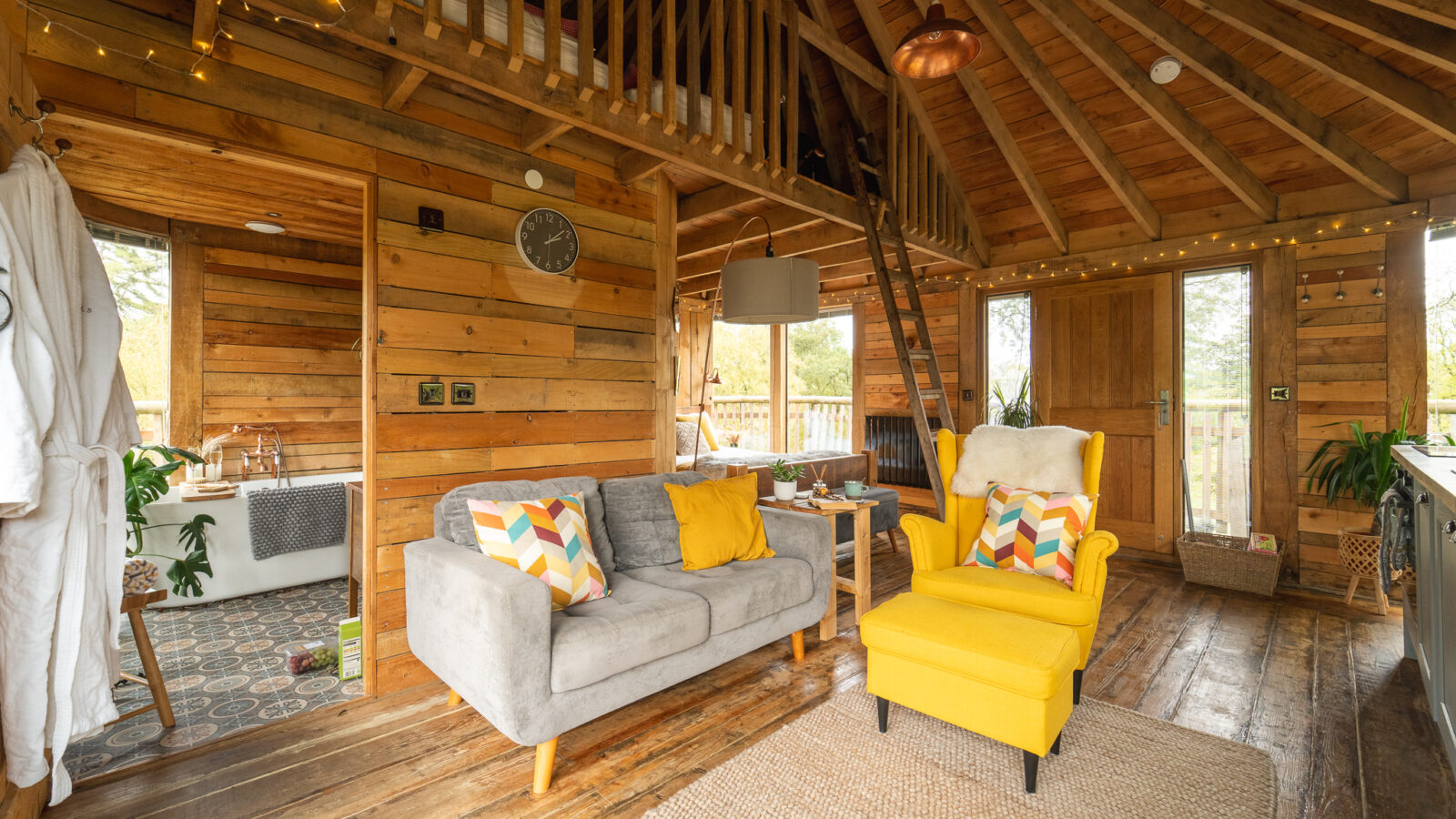 A cozy wooden cabin interior in Trawscwm features a gray sofa, yellow armchair, loft area, and decorative lights, evoking the charm of a treehouse retreat.