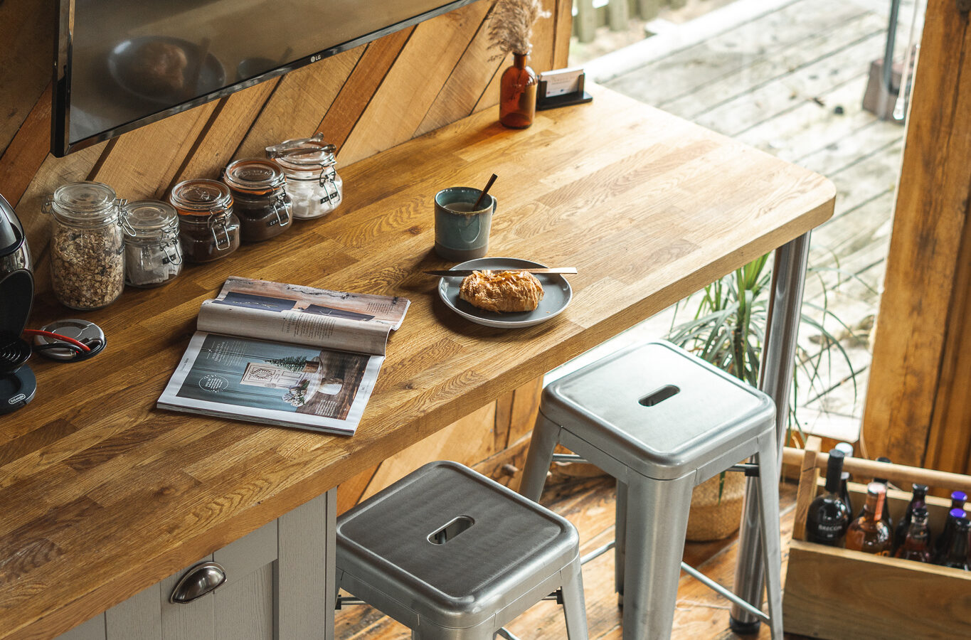 In the Trawscwm Treehouse, a modern kitchen nook features a wooden countertop, metal stools, and a TV overlooking the deck. Magazines and a pastry lie invitingly on the counter.