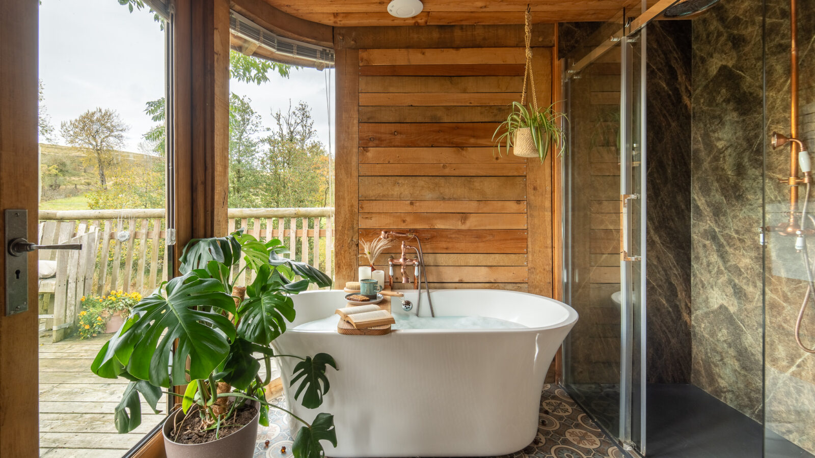 Experience a unique stay in this modern bathroom featuring a freestanding bathtub, shower, and large plant. Wooden walls and a window overlooking trees create the perfect Trawscwm Treehouse vibe, making it an exceptional treehouse accommodation getaway.