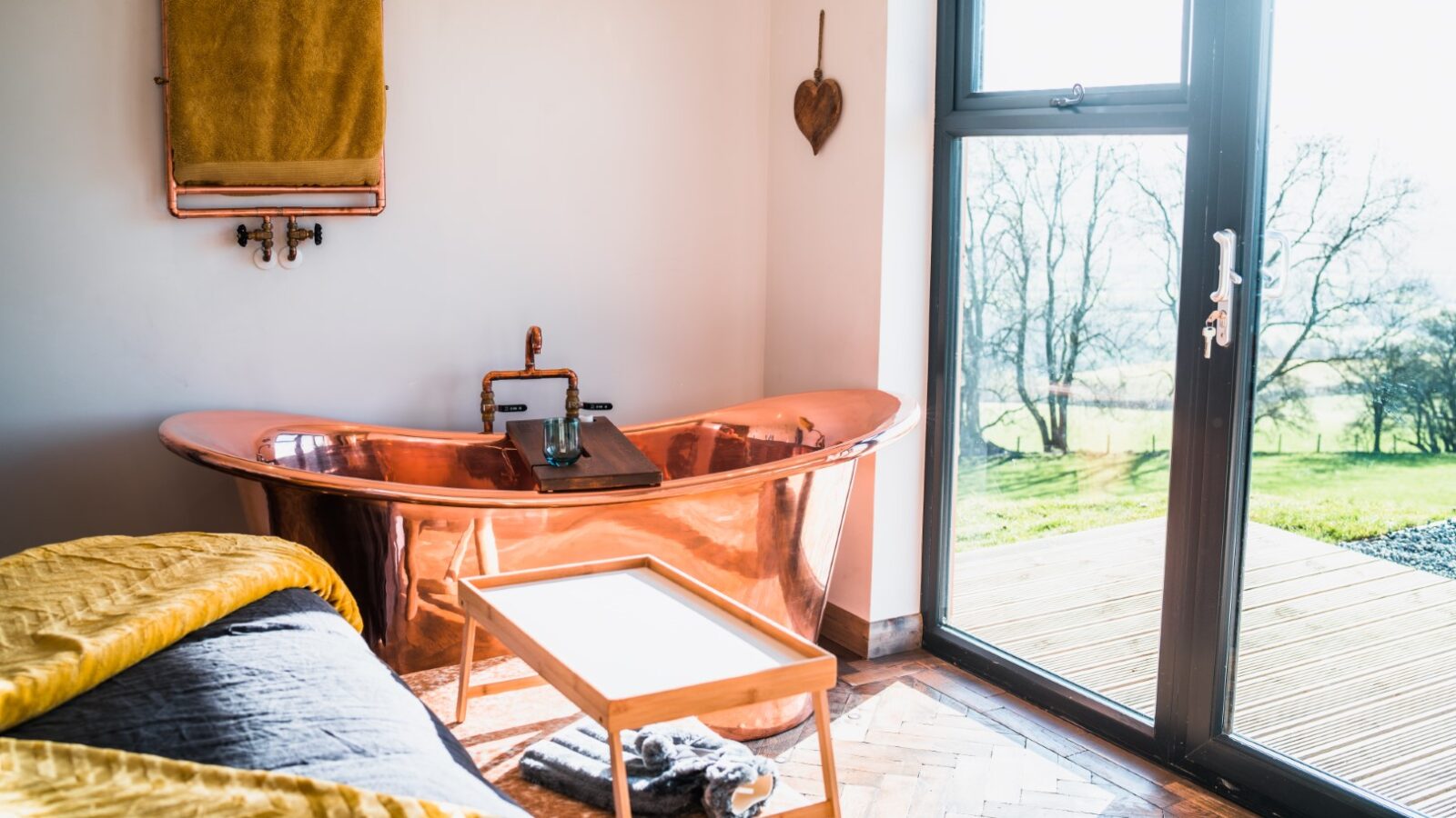 The bright bathroom at Penlan Lodges features a stunning copper bathtub, a yellow towel draped elegantly on a small table, and a large window that offers picturesque views of the surrounding greenery.