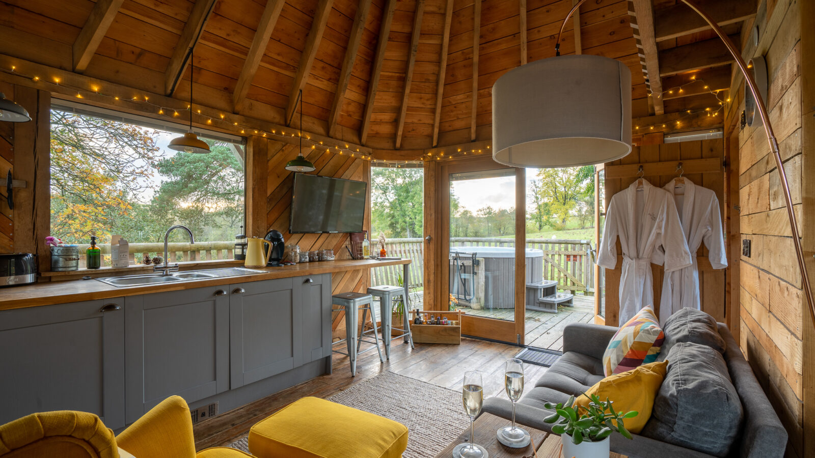 The Trawscwm Treehouse boasts a cozy wooden cabin interior, featuring a grey sofa and two yellow chairs. The inviting kitchen area and plush robes promise comfort, while the view of the hot tub outside adds an extra touch of luxury to this enchanting retreat.