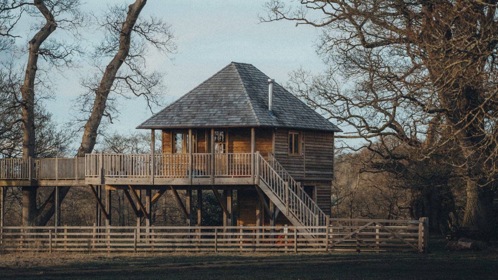 The Netherby Treehouse, with its wooden staircase and railing, stands elevated among leafless trees in a serene setting. The structure is surrounded by a wooden fence, embodying the charm of late autumn or early winter.