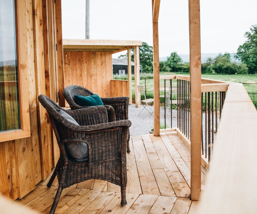 Two wicker chairs on a wooden porch offer a perfect retreat, overlooking a grassy landscape with a clear sky in the background.