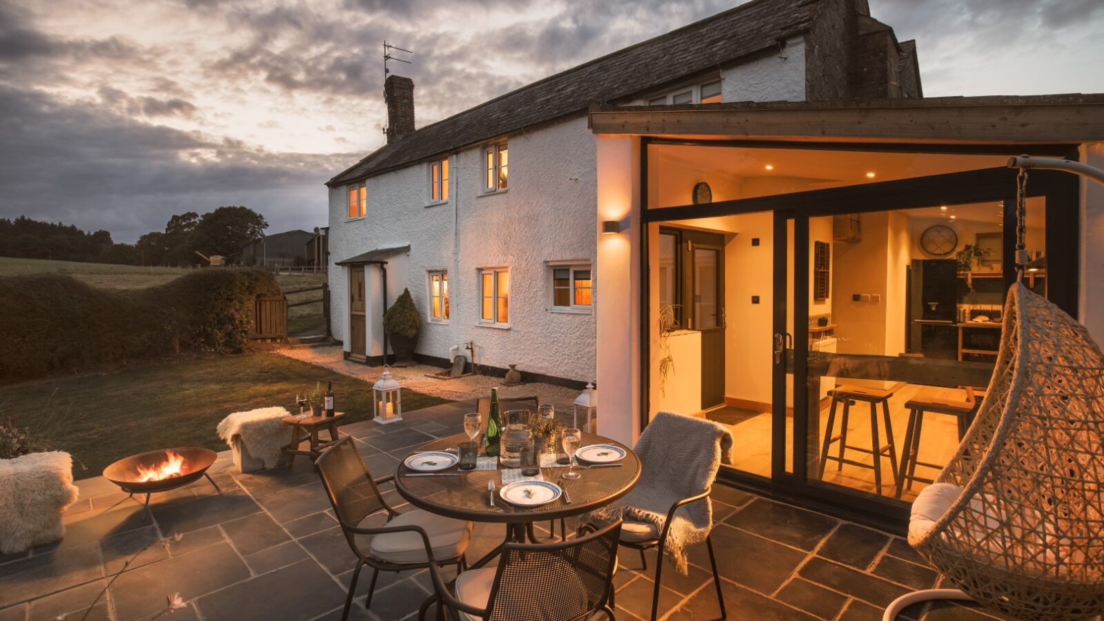 The outdoor patio at Fords Croft Cottages is enchanting at dusk, featuring a dining table set for four and a cozy fire pit nearby, offering a stunning view into the warmly lit house interior.