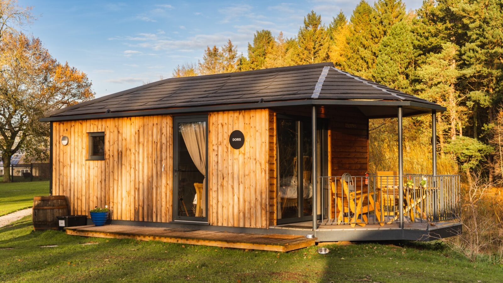 A charming small wooden cabin with a porch sits peacefully by the riverside, nestled in a grassy area surrounded by trees under a clear blue sky.