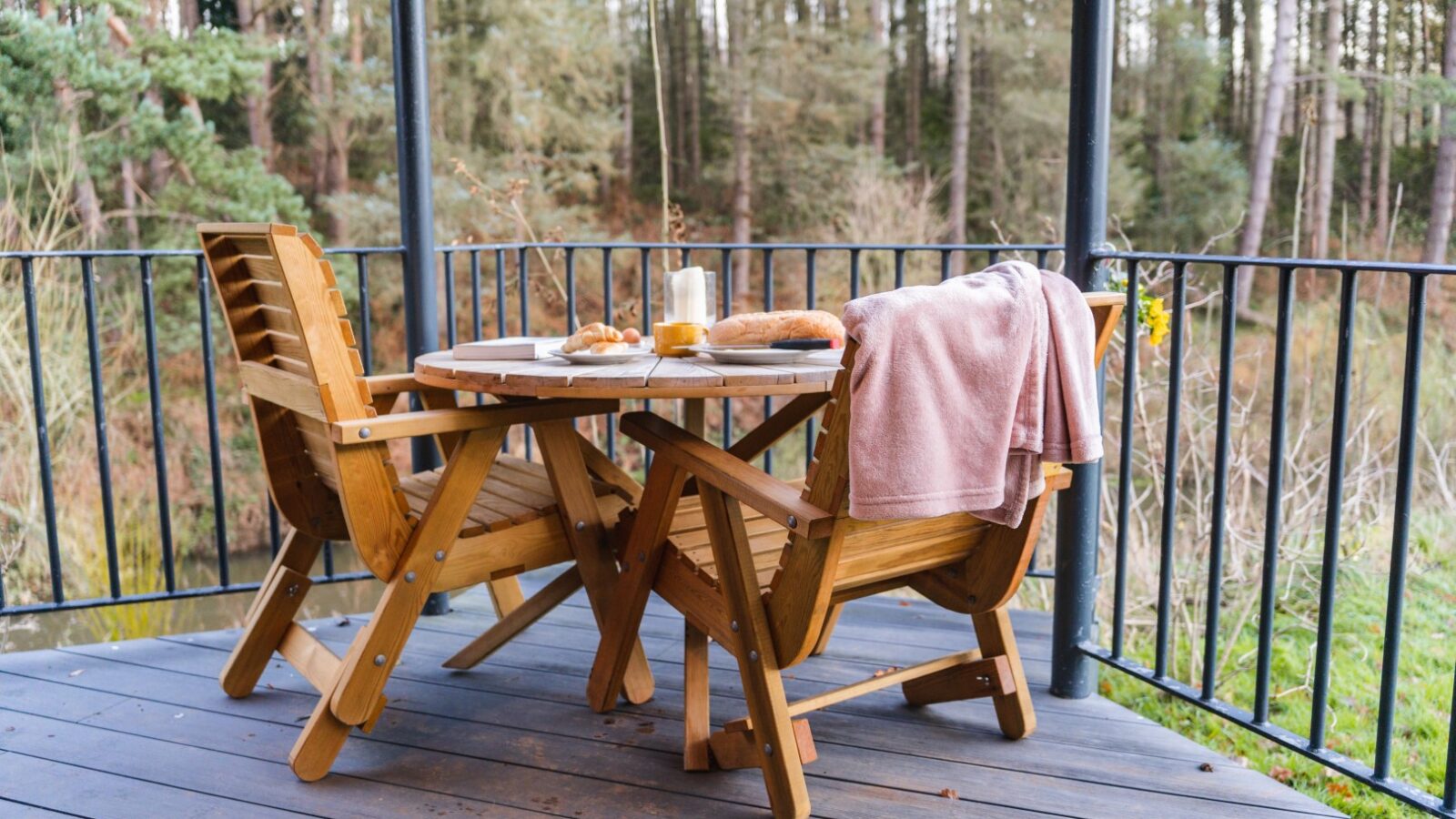 Wooden patio set with a round table and two chairs on a deck, featuring a pink blanket, mug, candle, and bread overlooking riverside cabins among the trees.