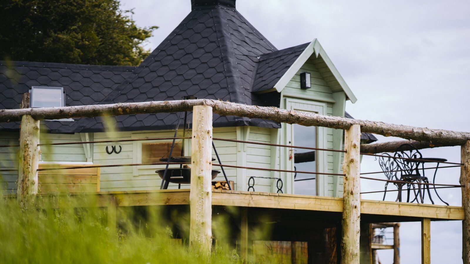 A small wooden cabin with a slate roof and enclosed porch sits on a grassy hill near the legendary Dragons Gate, offering outdoor furniture to enjoy the serene surroundings.