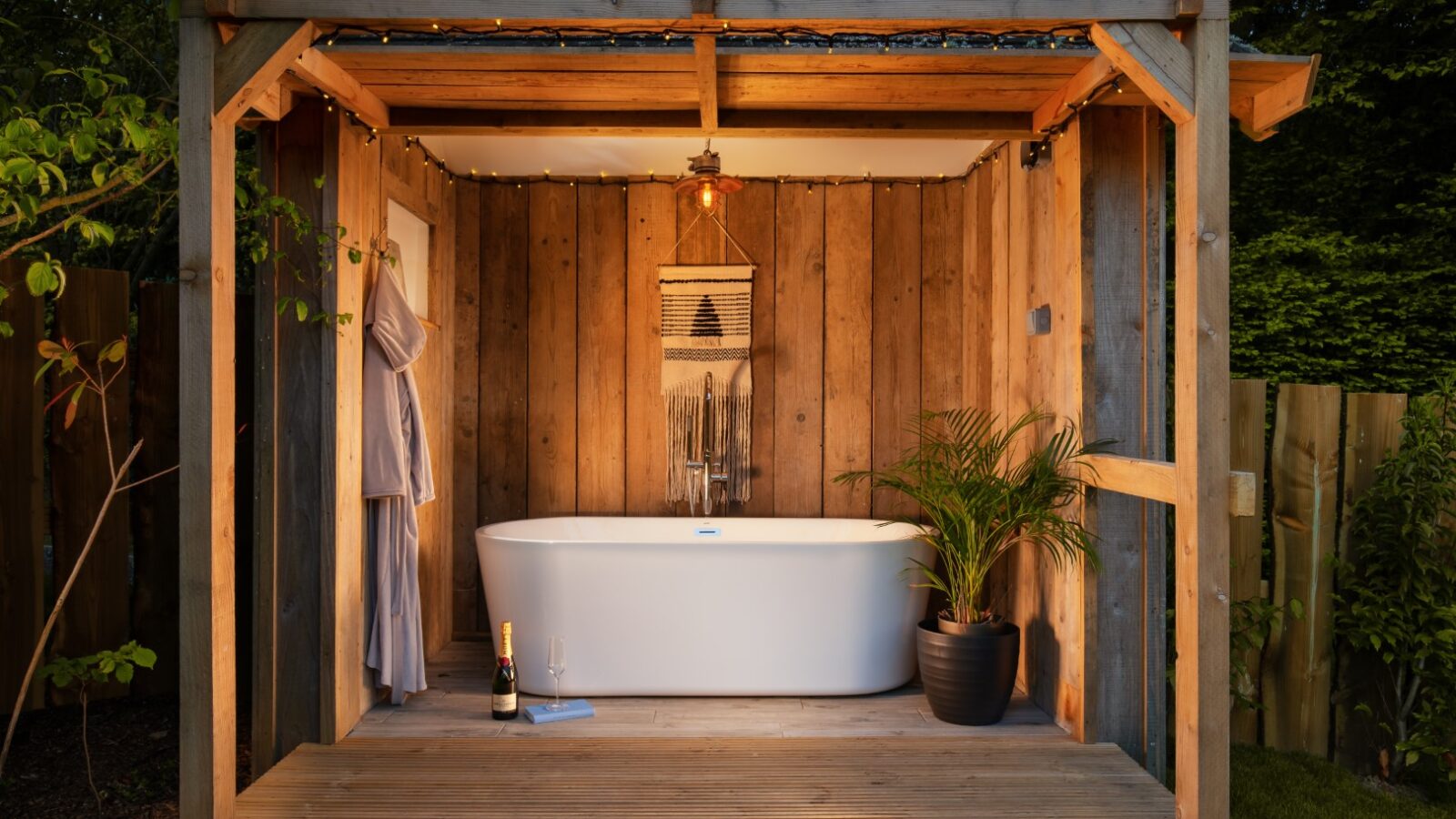 Nestled amidst nature, the outdoor wooden bathhouse features a freestanding tub, potted plant, and hanging lantern. A hidden gem for relaxation, it also includes a bottle of champagne on the floor to complete this serene escape.