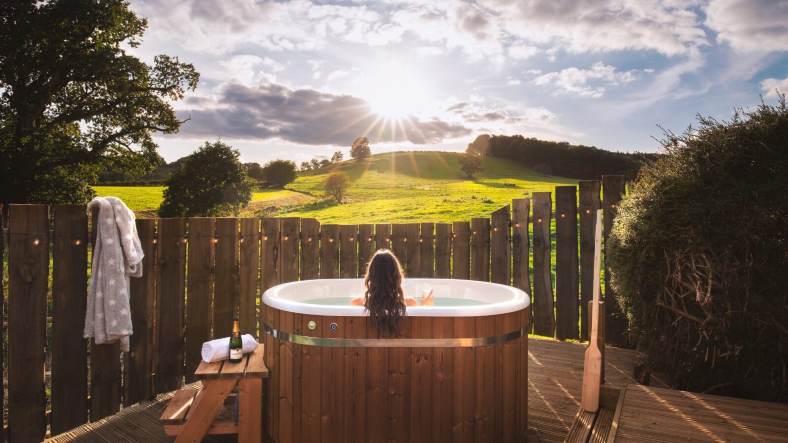 A person relaxes in a wooden hot tub outdoors at Fords Croft Cottages, overlooking a scenic countryside landscape under a partly cloudy sky.