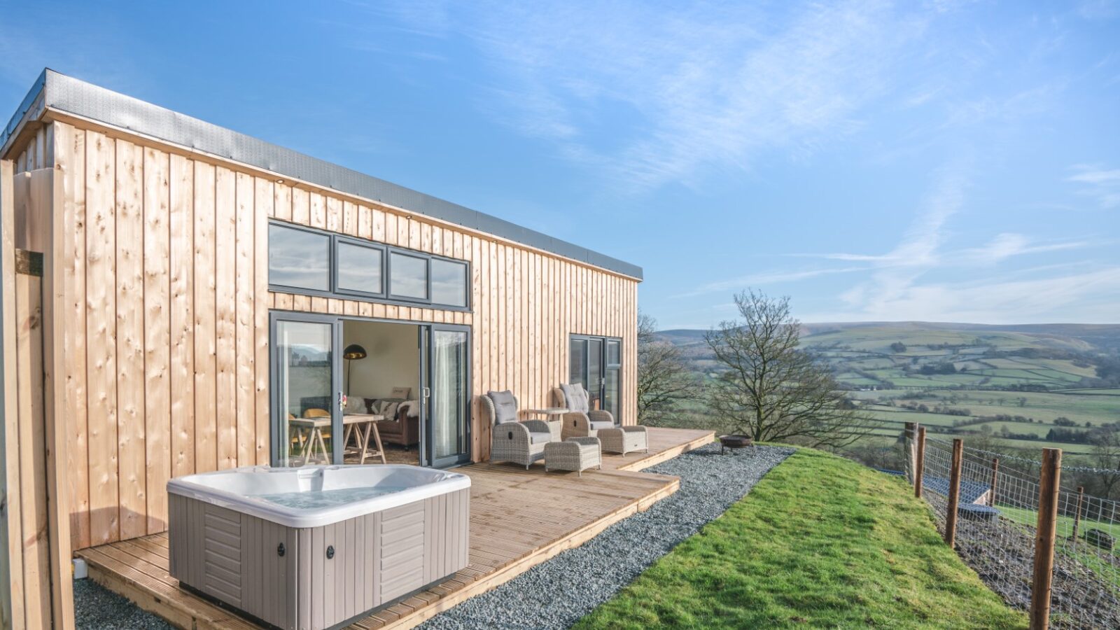 Penlan Lodges offer a modern wood cabin with a deck, outdoor seating, and a hot tub that overlooks a vast green landscape and hills under a clear sky—a perfect retreat for those seeking exceptional accommodation.