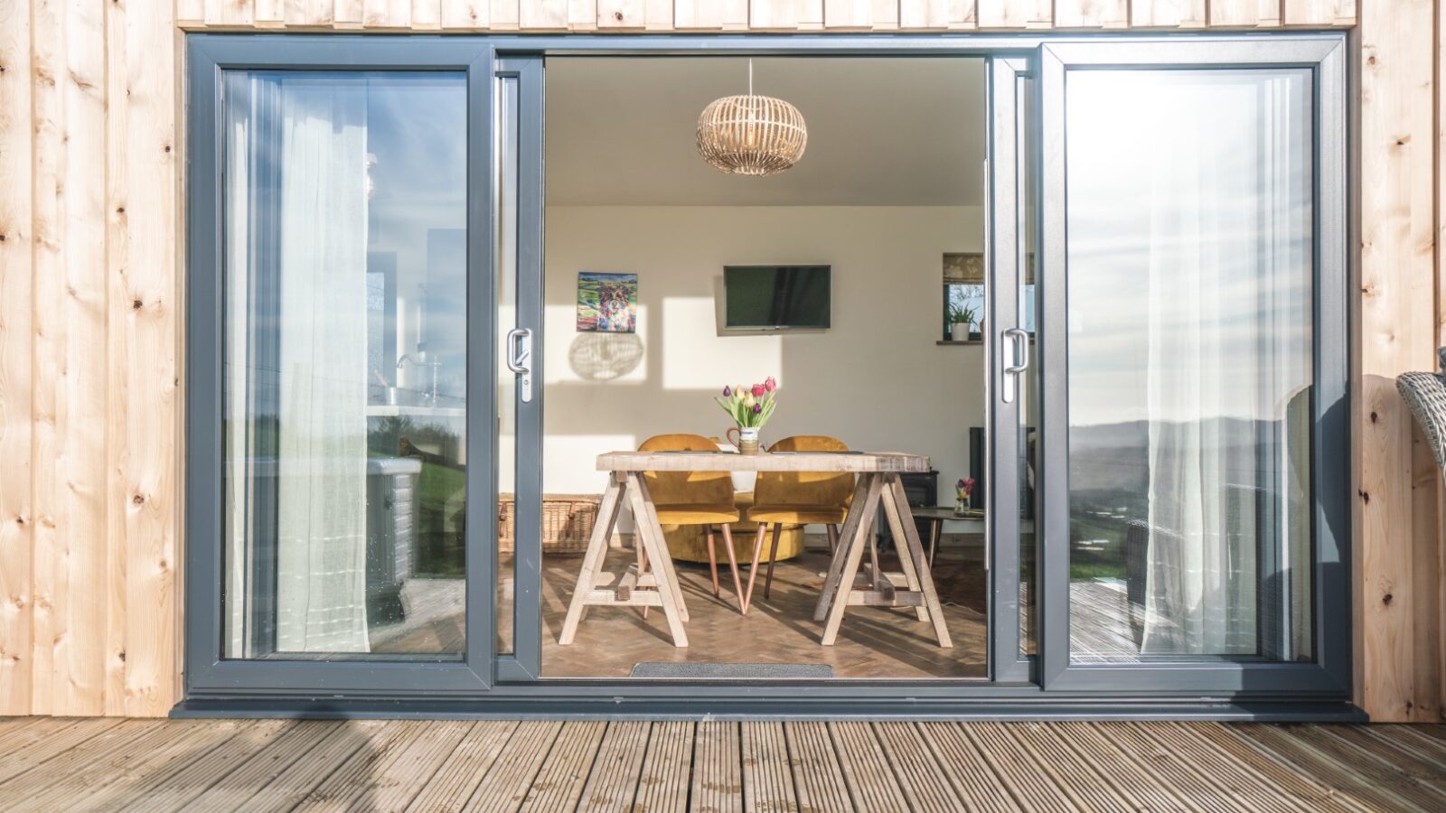 Sliding glass doors open to a wooden-floored room with a table, chairs, and wall-mounted TV, overlooking the sunny deck at Penlan Lodges.