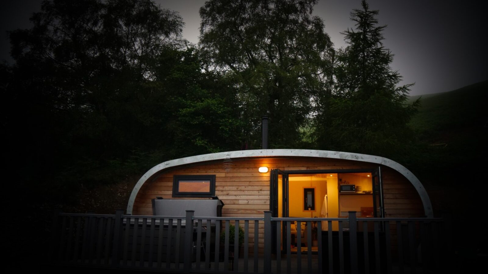 A small, illuminated wooden cabin with a curved roof and outdoor deck, set against a backdrop of trees at dusk, offers the ultimate glamping experience. Embrace nature with all the comforts found in luxury pods like Beacon View Pods.