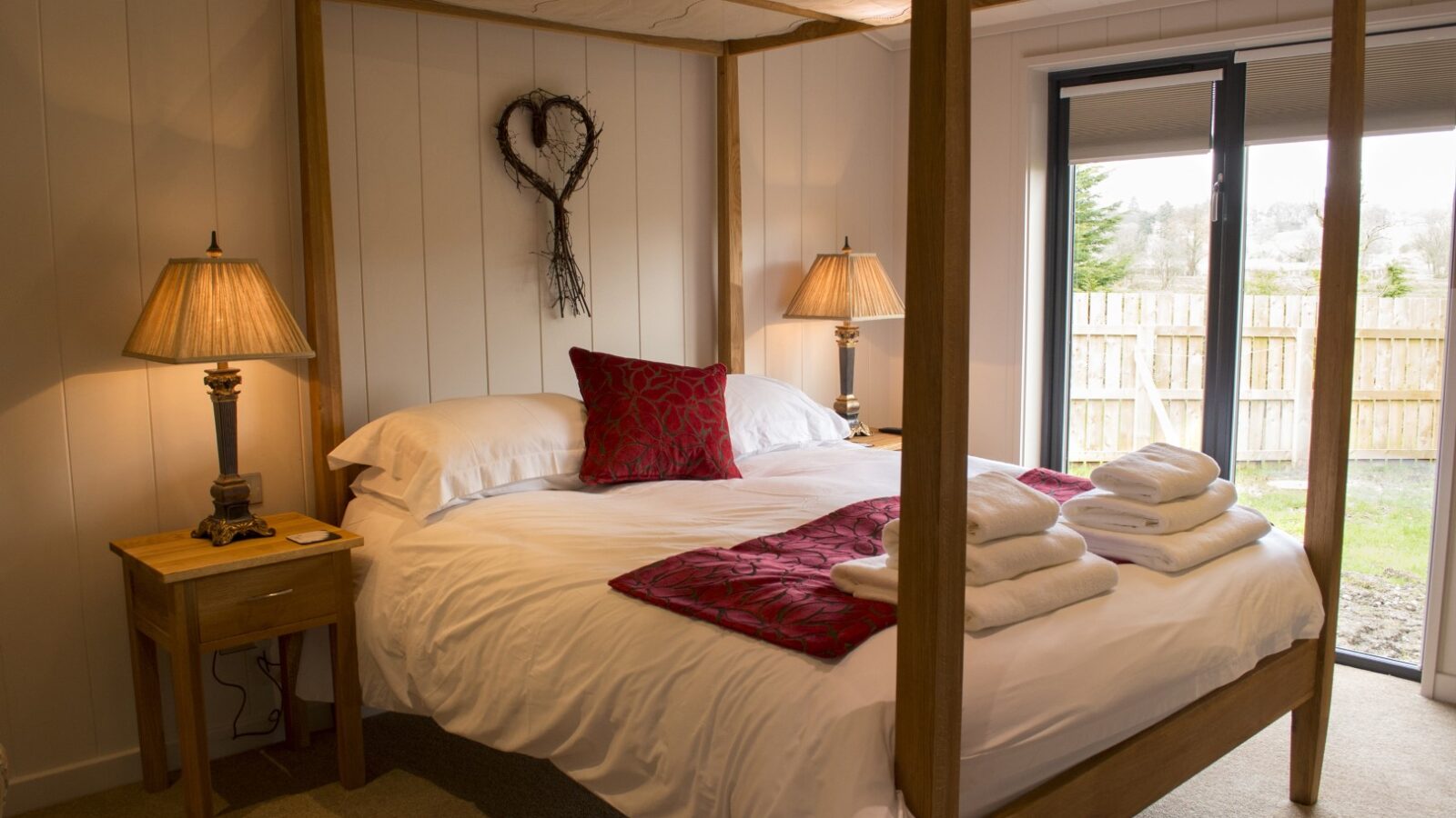 A cozy bedroom in a charming lodge features a wooden four-poster bed adorned with white bedding and red cushions. Two wooden bedside tables hold matching lamps. Towels are folded neatly on the bed, and the room offers a large window view of a fenced garden, reminiscent of serene lakeside retreats.