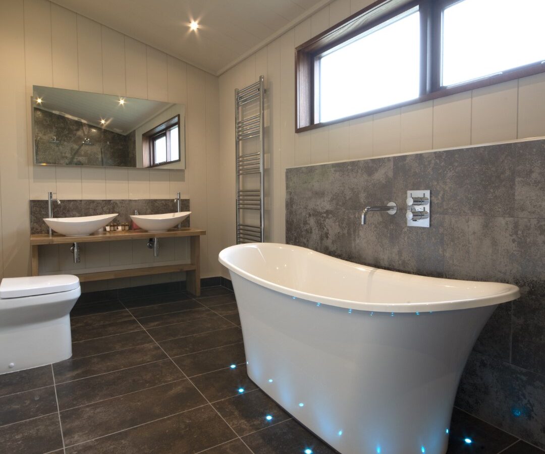 This modern bathroom, perfect for lakeside lodges, features a freestanding bathtub under a window, twin vessel sinks on a wooden vanity with a large mirror, a towel radiator, and a toilet. Gray tiles cover the floor and wall, creating a sleek, contemporary look.