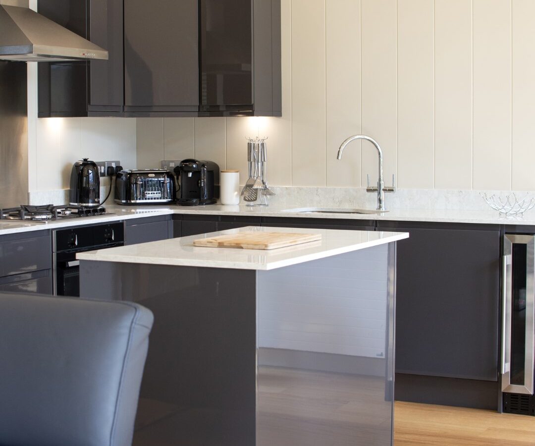 Modern kitchen with dark cabinets and stainless steel appliances exuding a lodge-like charm. A central island, topped with a wooden cutting board, complements the sleek sink with a curved faucet. Light wooden flooring flows beneath, while a black chair is partially visible in the foreground.