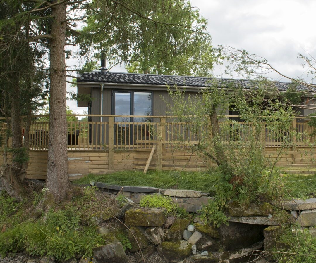 A modern cabin with large windows and a wooden deck, nestled among lush trees like a hidden lodge. A small stream flows in the foreground, adding to the serene forest setting reminiscent of tranquil lakeside retreats.