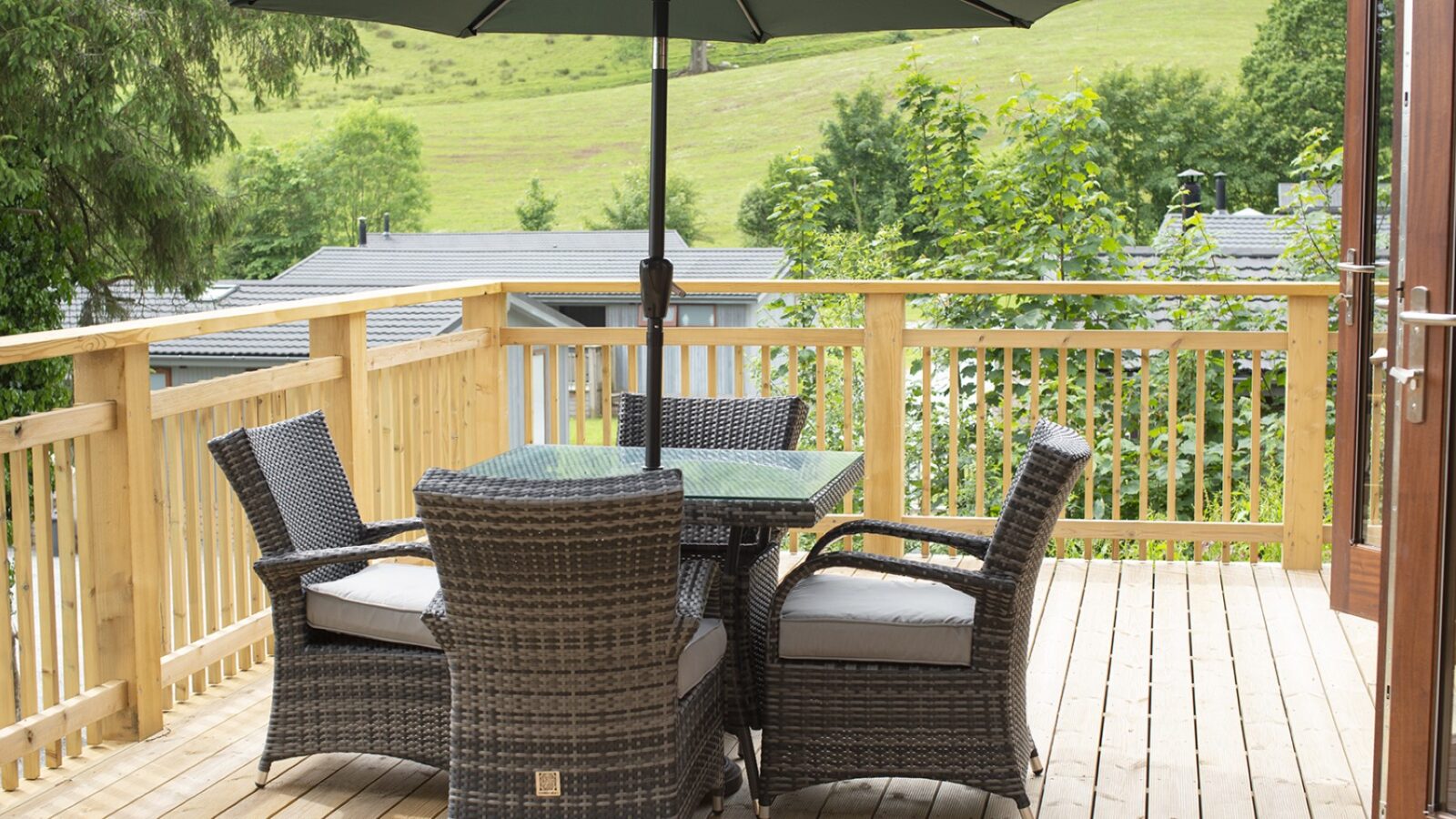 A wooden deck showcases a wicker dining set with four chairs and a glass-top table under a green umbrella, reminiscent of cozy lodges. The backdrop features lush greenery and distant rooftops, while a grassy hill rises beyond, evoking the serene charm often found by tranquil lakes.