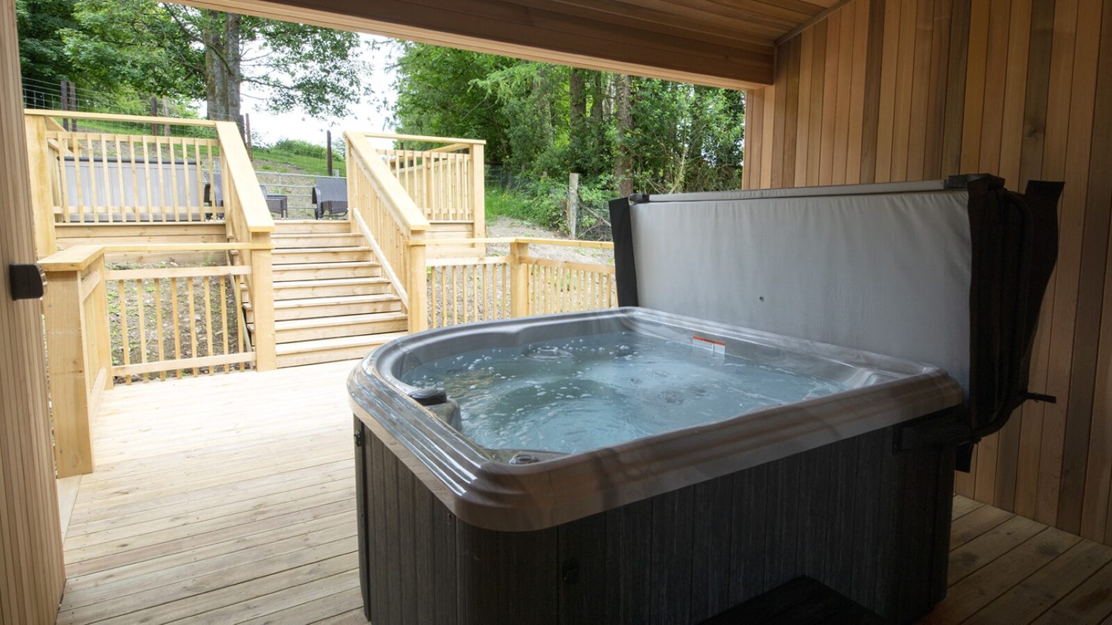 A serene outdoor hot tub sits on a wooden deck under a covered area, with steps leading to a lush garden surrounded by trees. Perfect for those staying in nearby lodges, it offers a tranquil escape reminiscent of the soothing surroundings found near picturesque lakes.