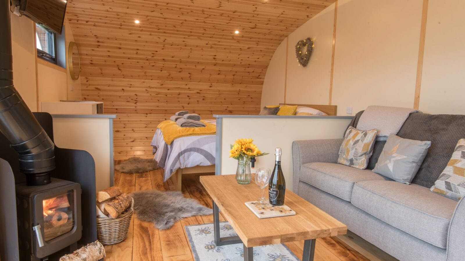 Cozy cabin interior with a wood-burning stove, sofa, coffee table, and bed. Decor includes sunflowers and logs near the stove, all enveloped in a charming setting reminiscent of Beacon View pods.