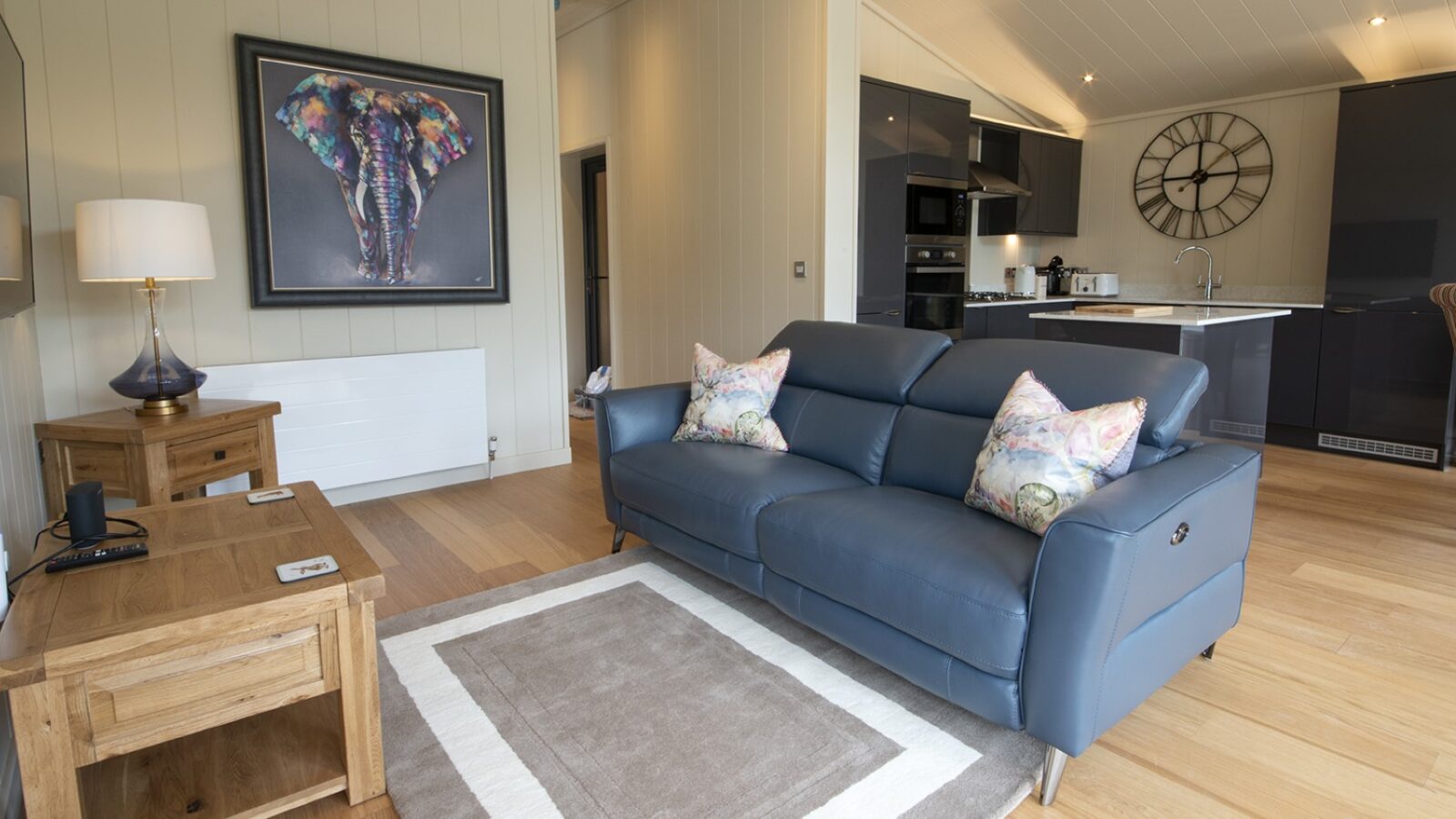 A modern living room with a blue leather sofa, colorful pillows, and a large framed elephant artwork exudes lodge-like charm. A wooden coffee table complements the aesthetic, and the kitchen area is visible in the background with dark cabinets and a large wall clock above the counter.