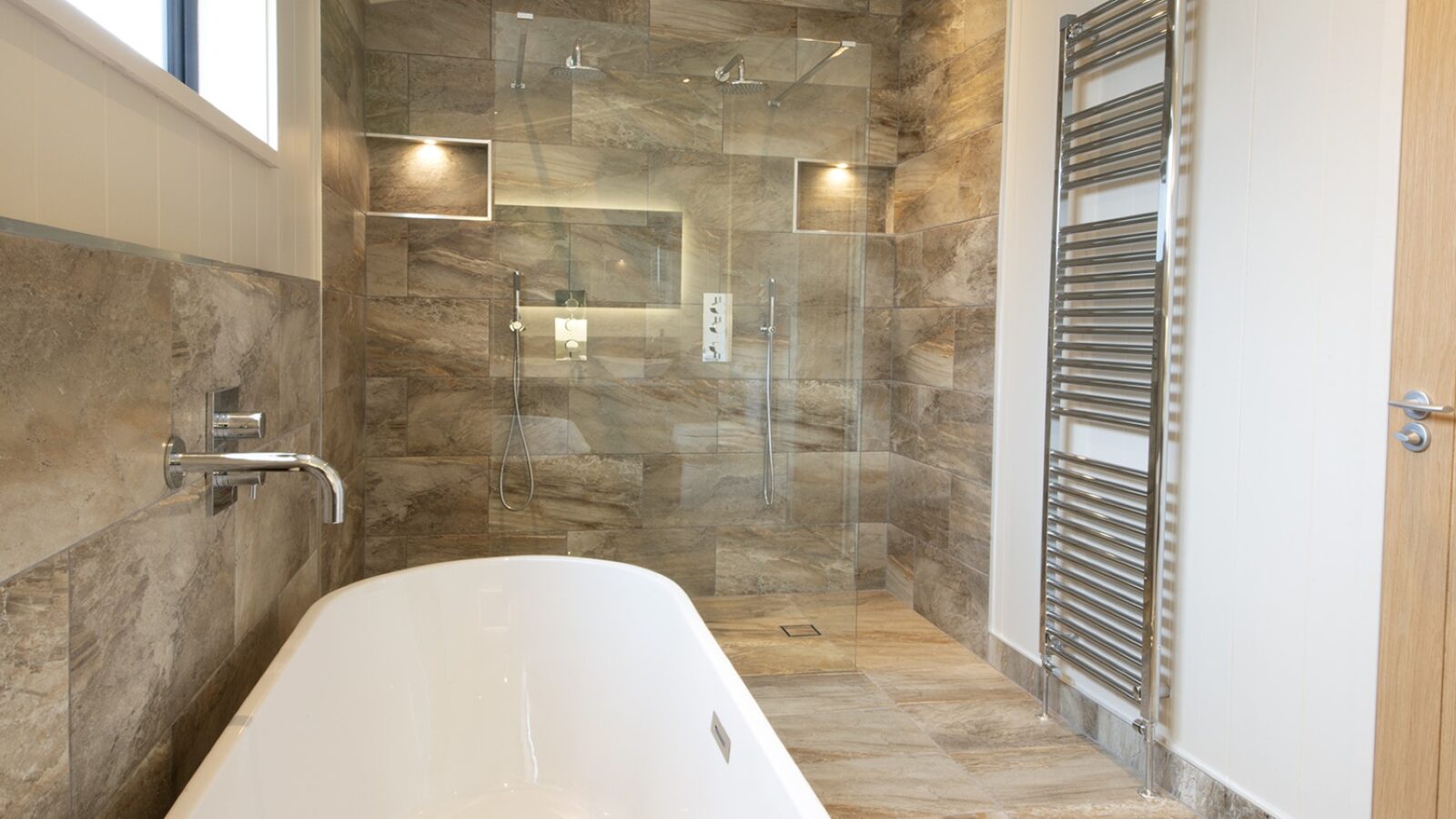 This modern bathroom, reminiscent of luxe lakeside lodges, features a large glass shower and a white freestanding bathtub. Brown stone tiles cover the floor and walls for a sleek, contemporary look. Natural light streams through a window on the left, illuminating the wall-mounted towel radiator.