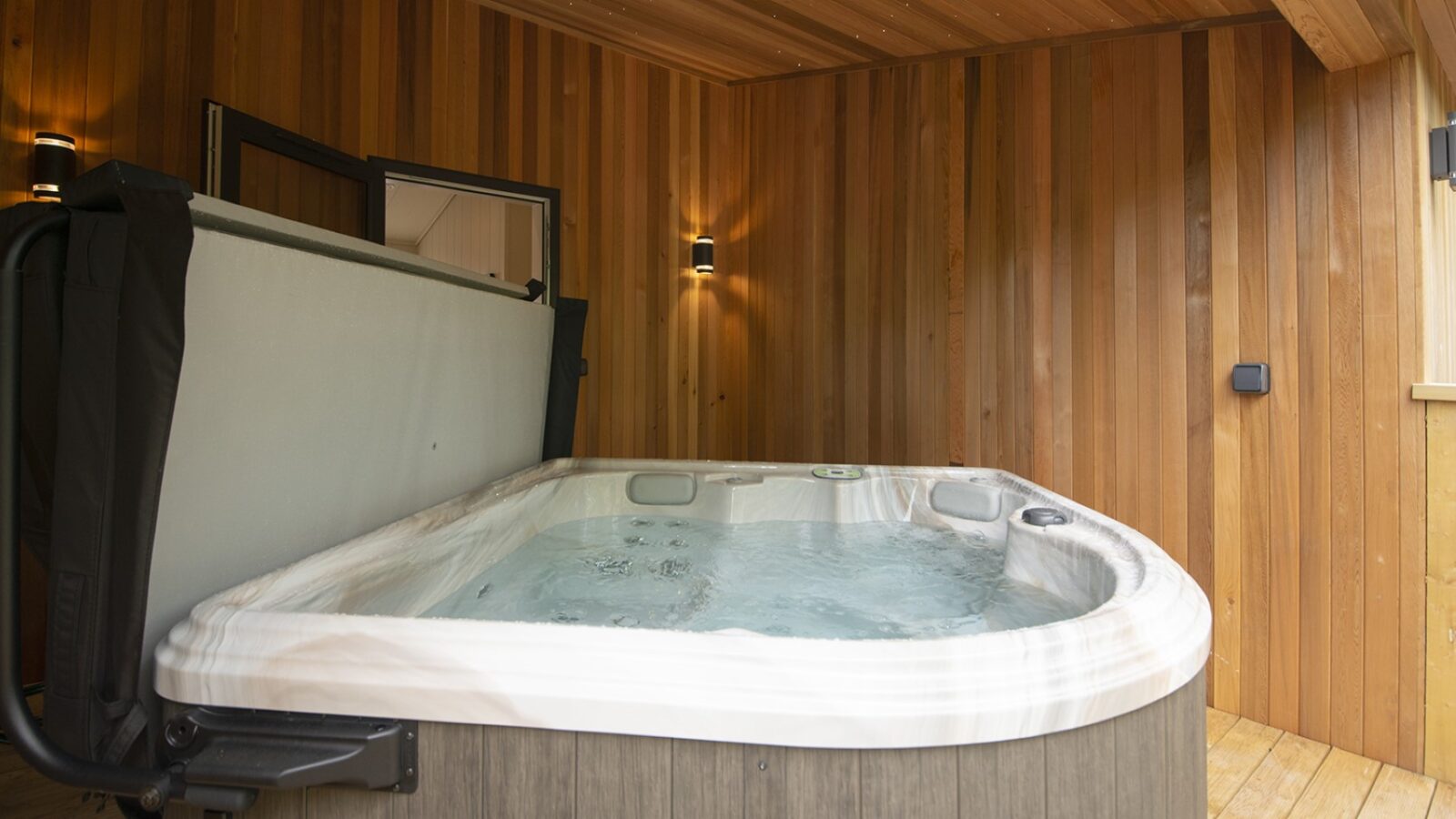 An indoor hot tub with swirling water graces a wooden-paneled room, echoing the cozy charm of lakeside lodges. Small wall lights add a warm glow, while a partially open cover rests on the left side, inviting relaxation.