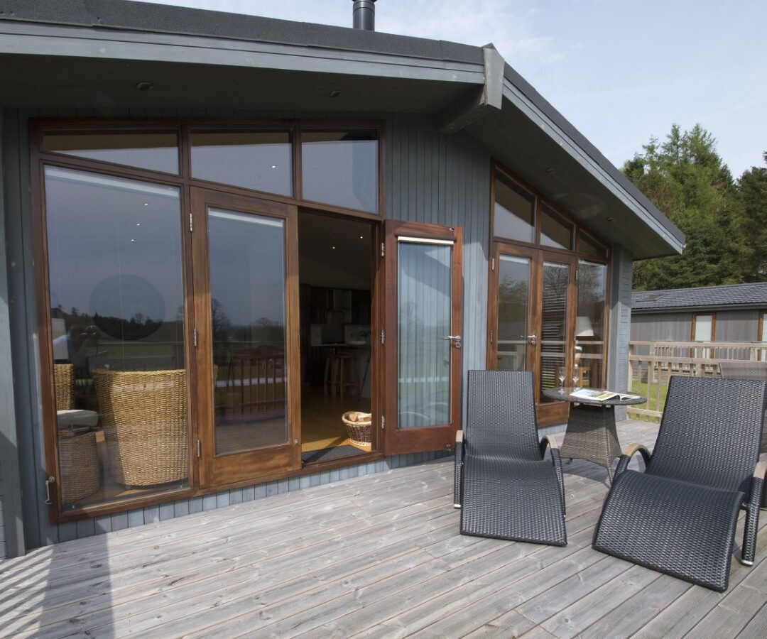 A wooden deck with two black lounge chairs overlooks a serene lake, complementing the modern house's large glass doors and windows. This lakeside lodge, with its sloped roof, is nestled amidst lush green trees and grass.