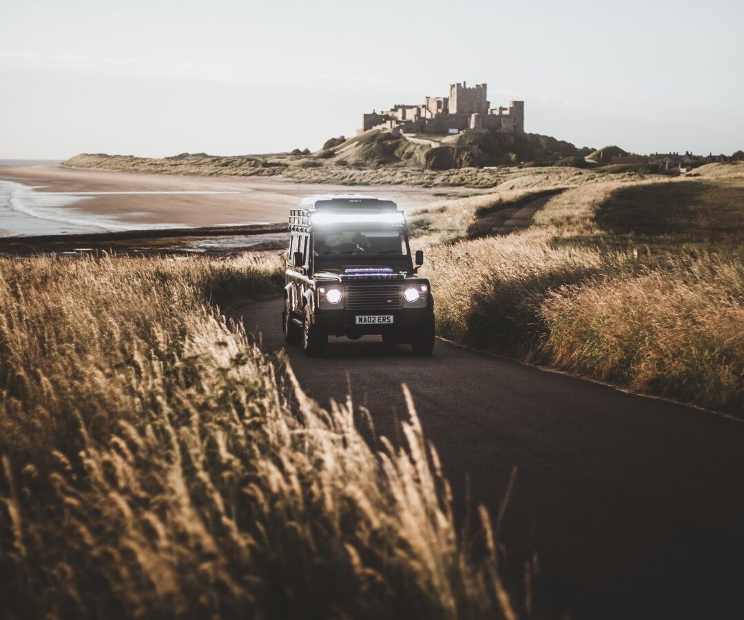 A black SUV, equipped with Defender Camping gear, drives on a narrow road through grassy fields, with a distant castle visible in the background.