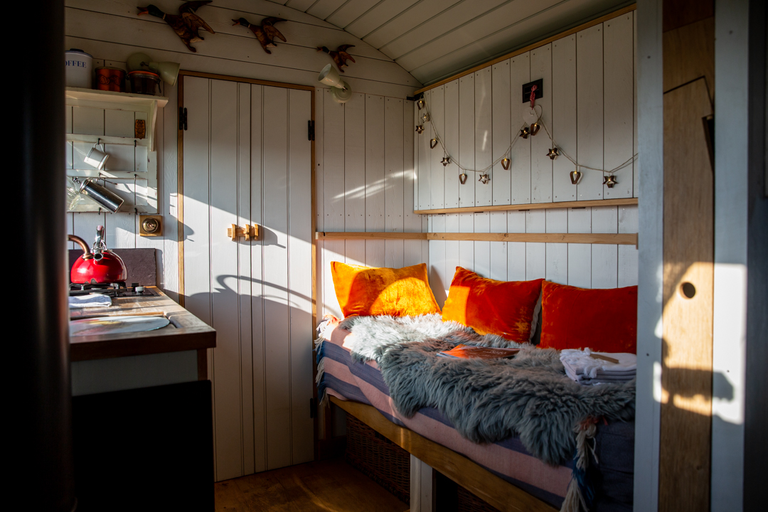 Experience the charm of glamping in this cozy cabin interior, complete with a small kitchen, a vibrant red kettle, and a bed adorned with plush orange cushions. The decorative lights on the wall add that extra touch of magic to your Dimpsey retreat.