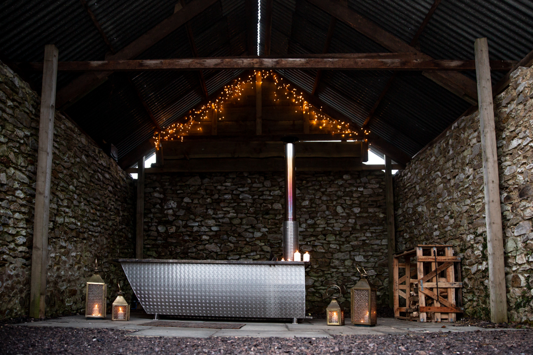 An outdoor metal bathtub under a rustic shelter with stone walls, surrounded by lanterns and string lights, captures the essence of Dimpsey glamping.