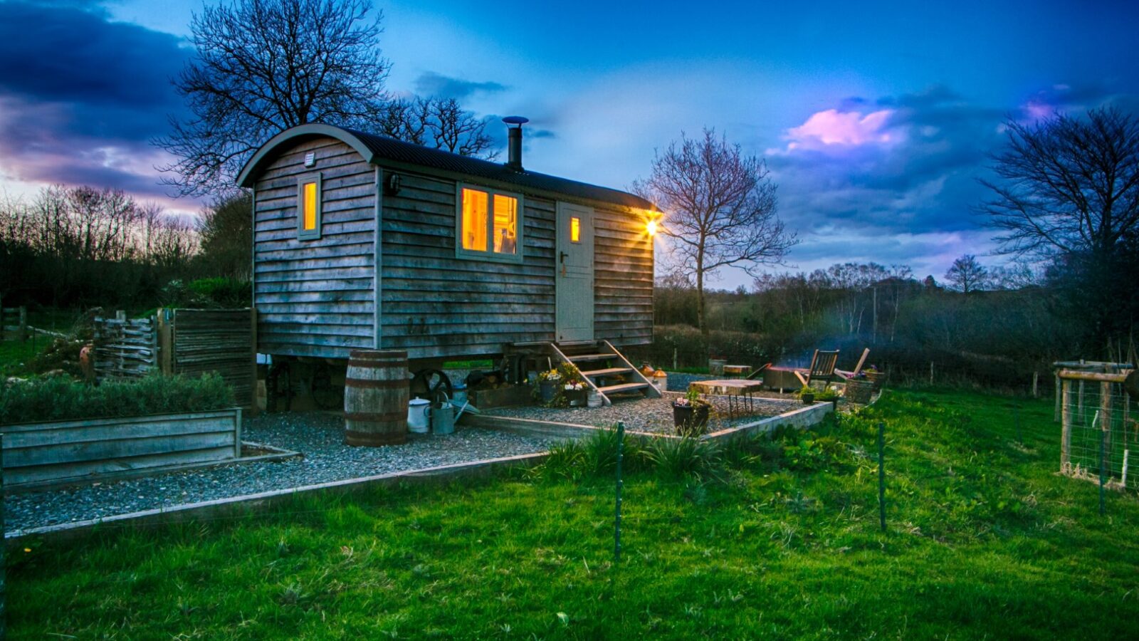 A cozy wooden cabin stands illuminated at twilight, offering a unique glamping experience amidst trees and a grassy landscape.