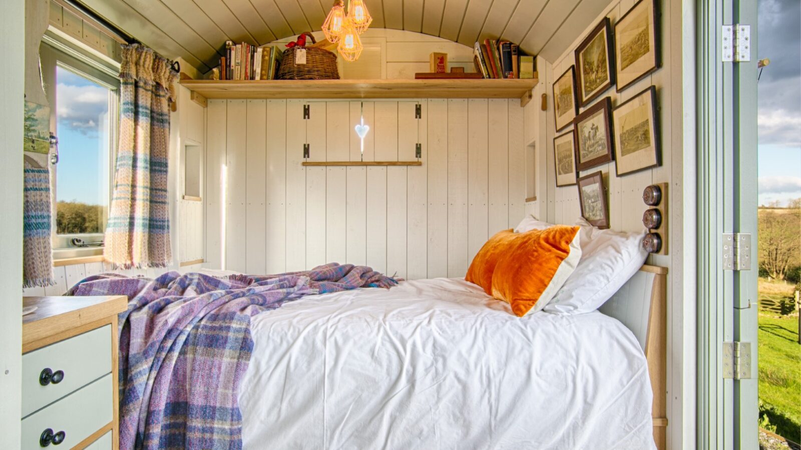 Cozy tiny home bedroom with white walls, a bed adorned with orange pillows, a shelf stacked with books, and windows draped in plaid curtains. Perfect for those seeking the charm of Glamping. Experience Dimpsey magic as you unwind in this quaint retreat.