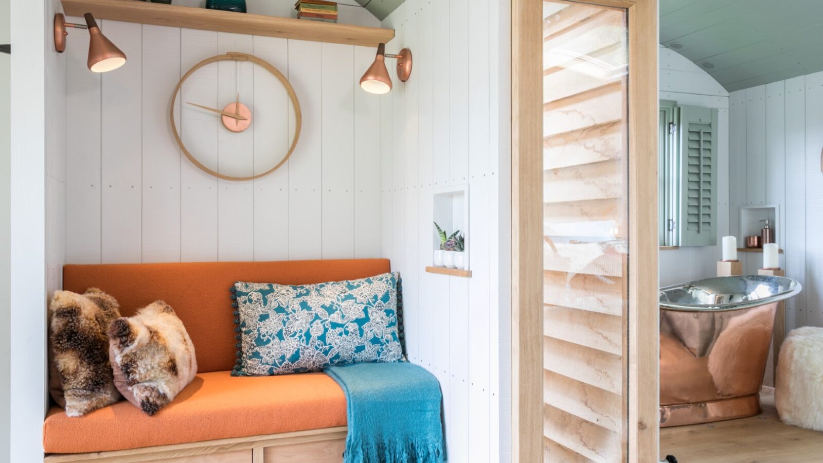 Cozy room with an orange sofa, decorative pillows, a round wall clock, and a copper bathtub in the corner—perfect for a dimpsey glamping retreat.