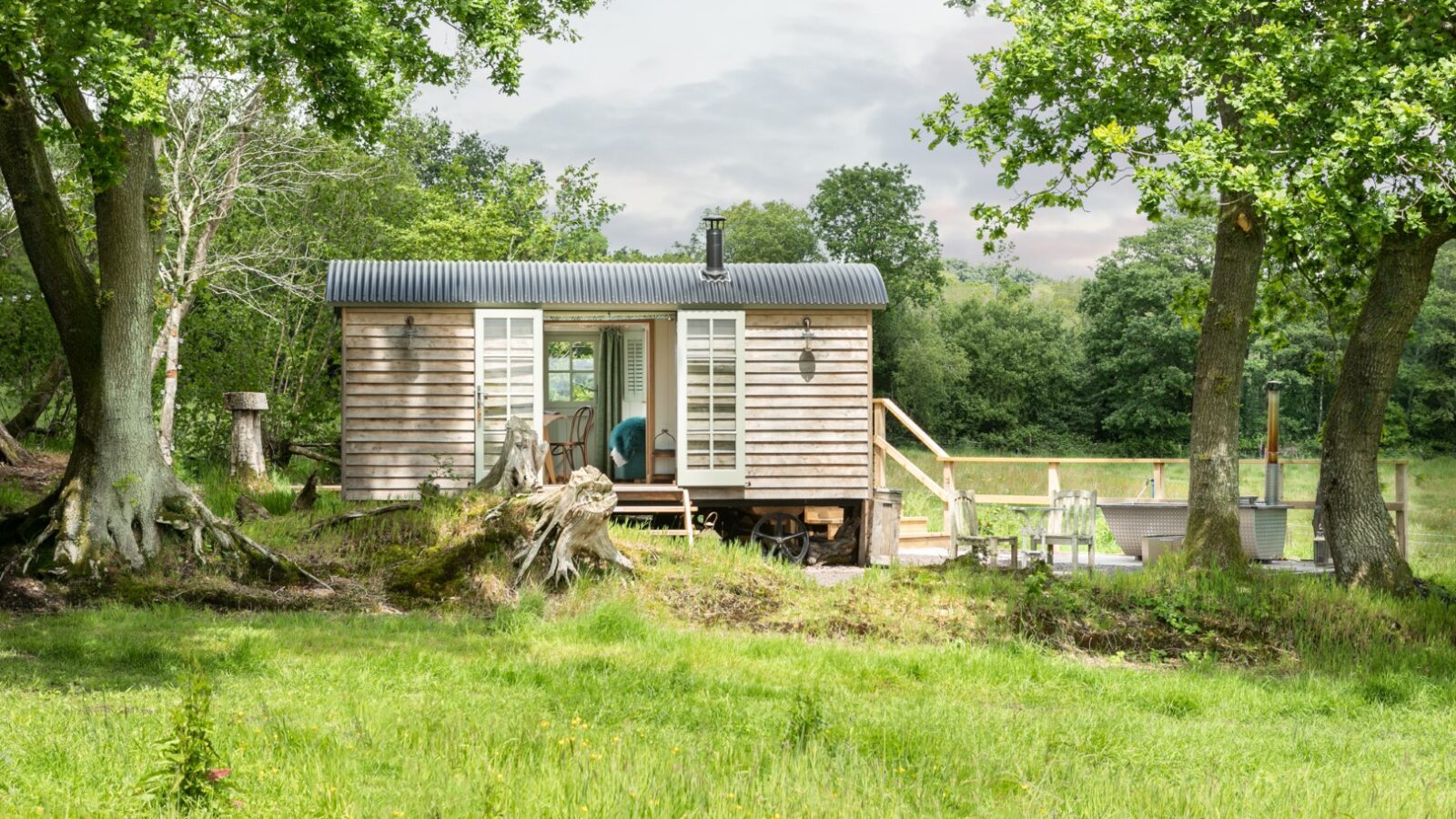 A charming wooden cabin with open doors rests in a grassy area, embraced by trees and a deck with seating, offering the perfect Dimpsey glamping escape.