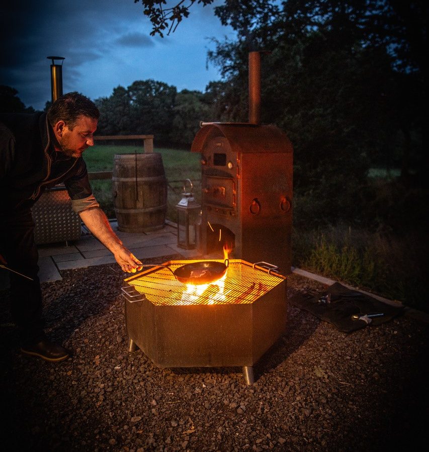 At Dimpsey, a person embraces glamping tranquility, tending to a fire in a metal pit beside a barrel and a wood-fired oven outdoors at dusk.