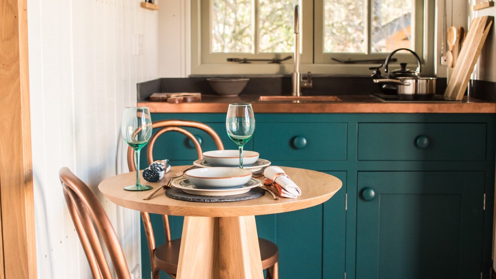 A cozy kitchen with teal cabinets, wooden chairs, and a round table set with plates, glasses, and cutlery near a window evokes the charm of glamping, blending rustic comfort and style.