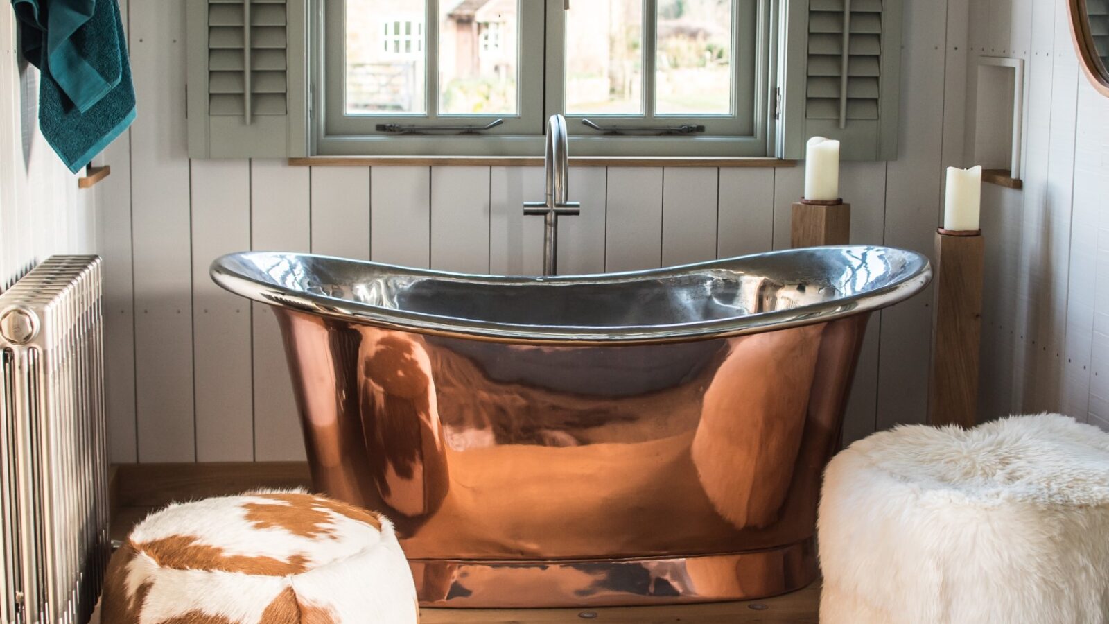 A copper bathtub sits in a cozy Dimpsey bathroom with shuttered blinds and rustic decor, including a cowhide stool and fluffy ottoman—perfect for a touch of glamping luxury.