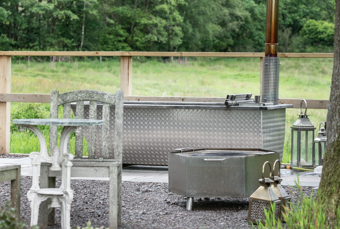 Experience the charm of glamping with an outdoor seating area featuring a metal hot tub, cozy benches, lanterns, and lush greenery in the background—perfect for a Dimpsey Glamping getaway.