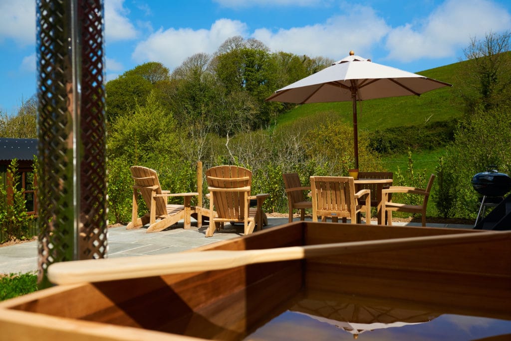 Discover tranquility at Dittisham Hideaway, where a serene outdoor patio with wooden chairs and a table rests under a large umbrella. Enjoy the foreground's charming wooden hot tub, surrounded by lush greenery and a grassy hill, all beneath a blue sky scattered with clouds.