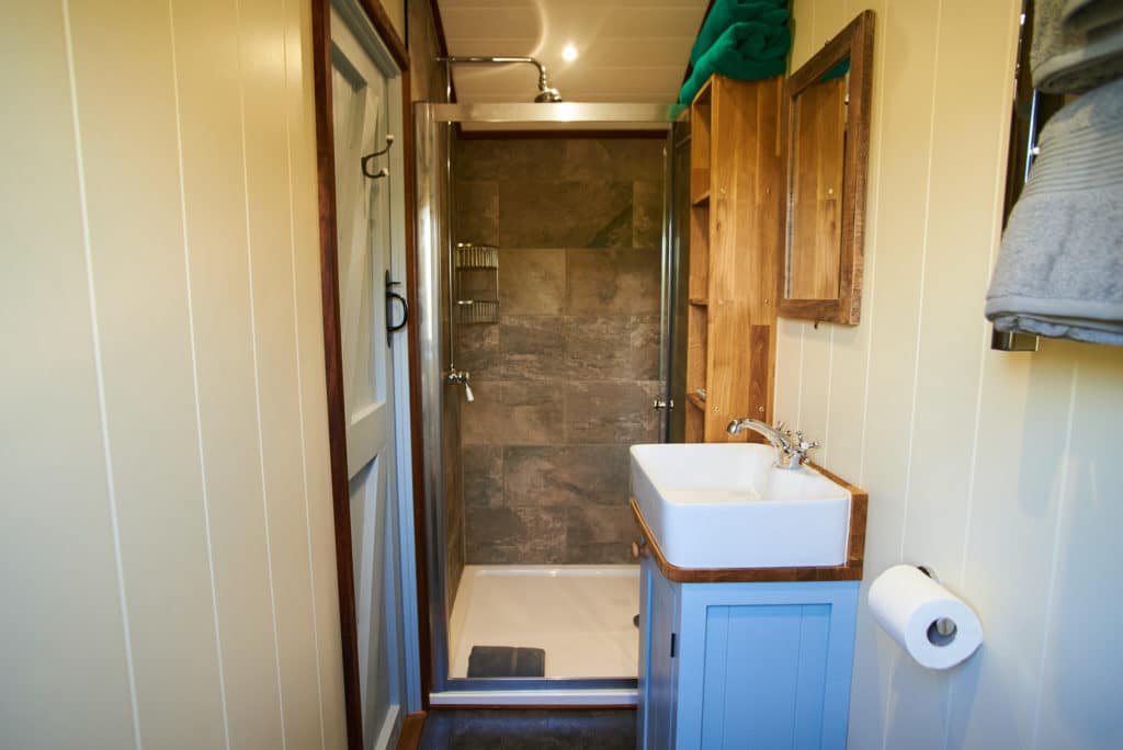 A small bathroom in the charming style of Shepherds Lodges features a shower with slate tiles, a rectangular sink on a blue vanity with a faucet, and a wooden-framed mirror. Cream-colored panel walls complement the space, complete with a towel shelf above and toilet paper holder beside.