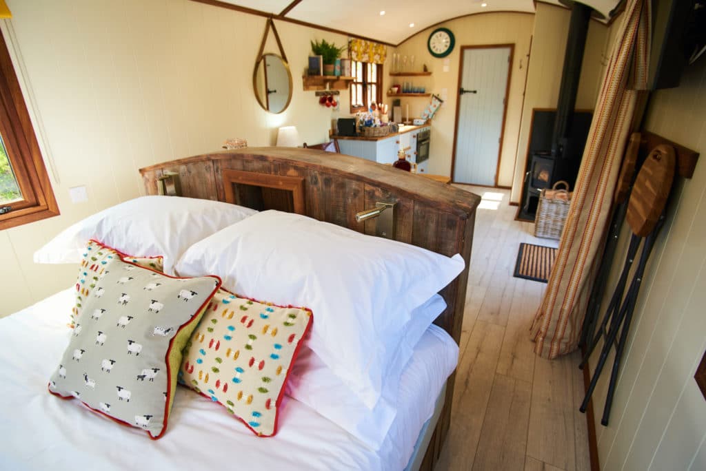 The cozy tiny cabin interior, reminiscent of a Dittisham Hideaway, features a neatly made bed with patterned pillows. A small kitchenette with wooden accents complements the space, while light-filled windows and a clock on the wall create a warm and inviting Shepherds Lodges ambiance.