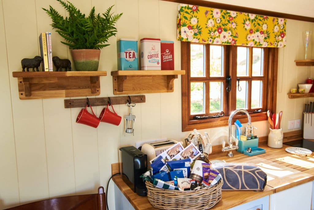 In a cozy kitchen corner reminiscent of a Dittisham Hideaway, a wicker basket brimming with snacks sits on a wooden counter. Above, two shelves hold tins labeled tea, coffee, and sugar. The floral curtains by the window and the hanging red mugs evoke the charm of Shepherds Lodges.