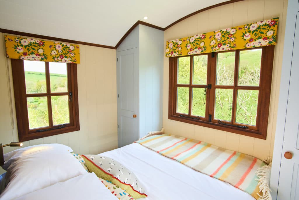 Cozy bedroom interior in Dittisham's Shepherds Lodges, featuring a double bed adorned with a striped blanket and floral cushions. Two windows with charming floral-patterned curtains reveal the serene green landscape outside. Light-colored walls and a closed cabinet complete this hideaway.